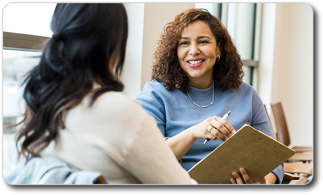 A curly-haired healthcare provider holding a clipboard smiles while speaking with a patient seated across from her, seen from behind.